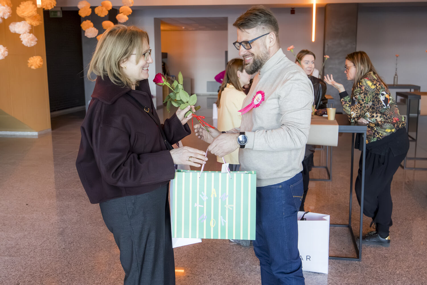 Mężczyzna wręcza Mamie kwiat i prezent, oboje się uśmiechają. W tle inne osoby rozmawiają. To moment finałowy spotkania „Oddech dla Mam” z okazji Dnia Kobiet w Solar.